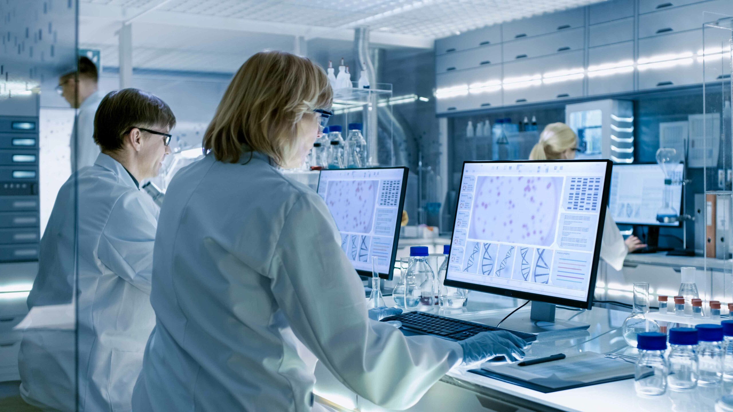 Female and Male Scientists Working on their Computers In Big Modern Laboratory. Various Shelves with Beakers, Chemicals and Different Technical Equipment is Visible.